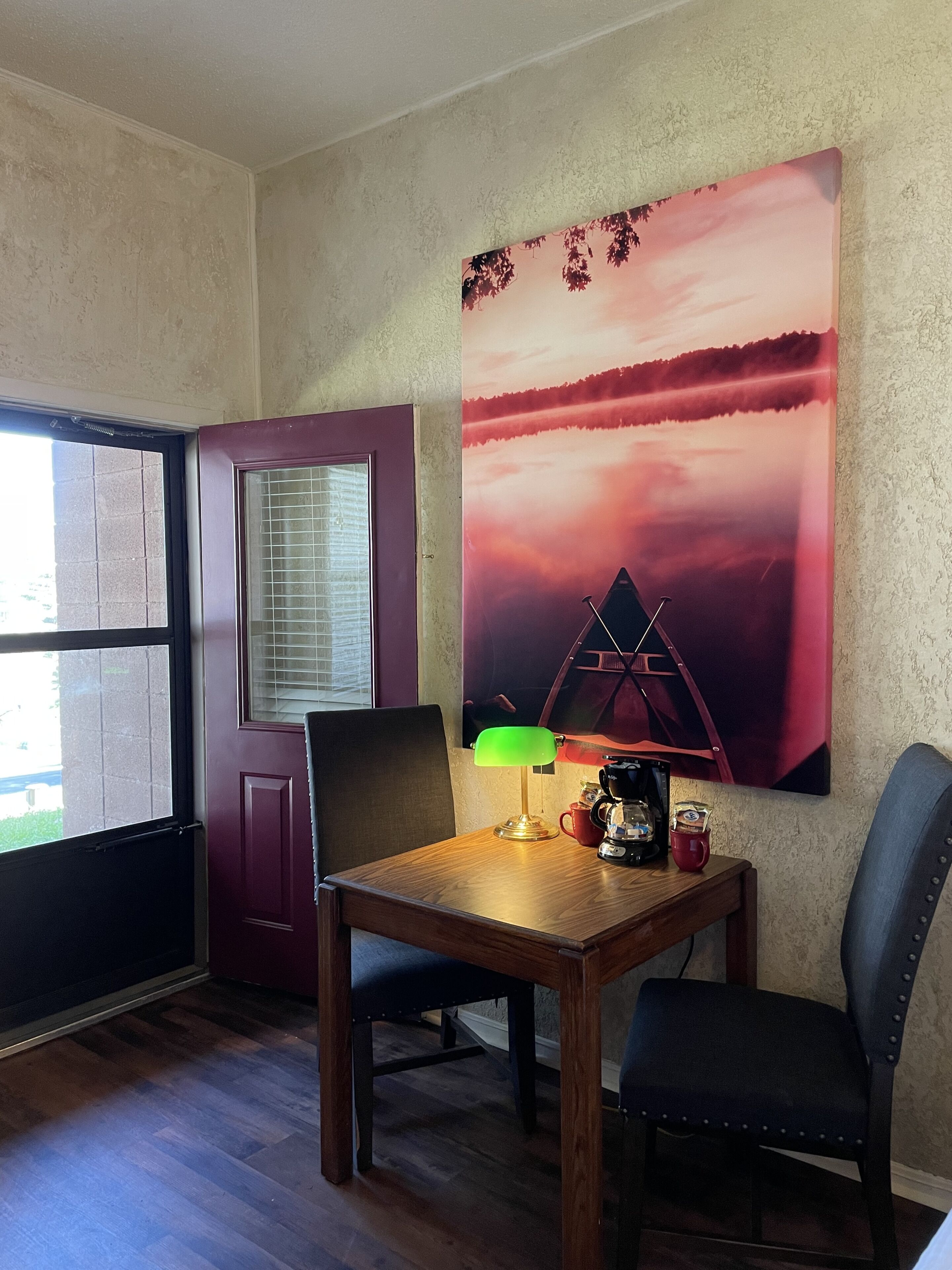 A room with a wooden table, two chairs, a lamp, a coffee maker, and a large painting of a sunset.