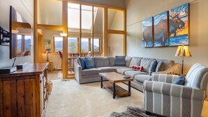 A modern living room with a sectional sofa, a wooden coffee table, and a striped armchair. There is a painting of moose on the wall, a lamp, and a view of mountains through large windows. - 2056 The Pines 2 Bedroom Condo by RedAwning (Keystone)
