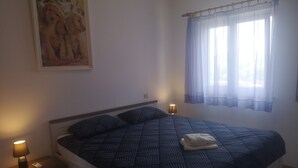 Un dormitorio con cama, una mesita de noche con lámpara, un cuadro de perros y una ventana con cortinas traslúcidas.