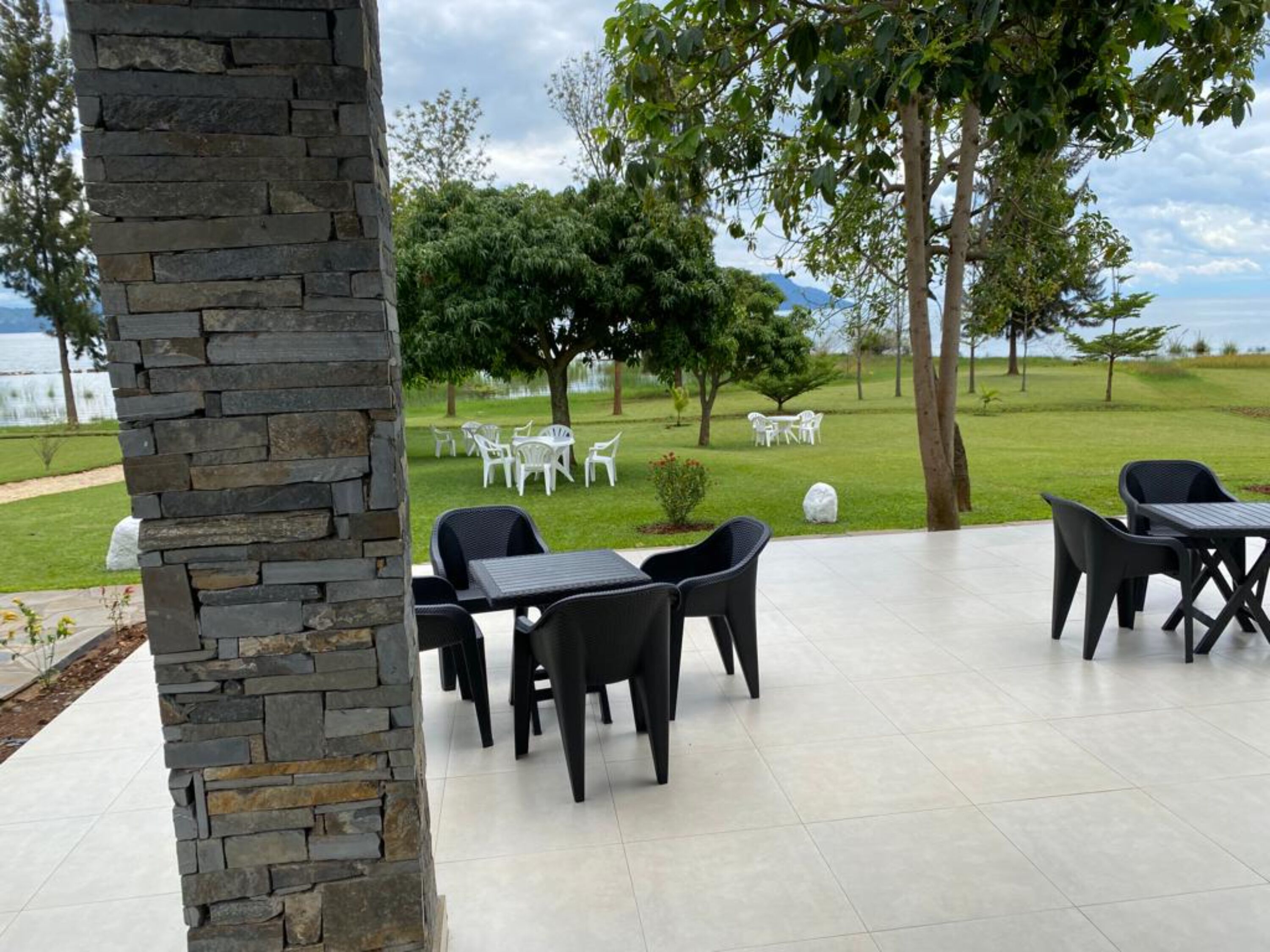 An outdoor patio area with stone walls, black chairs, and a view of a grassy landscape with trees and a body of water in the distance.