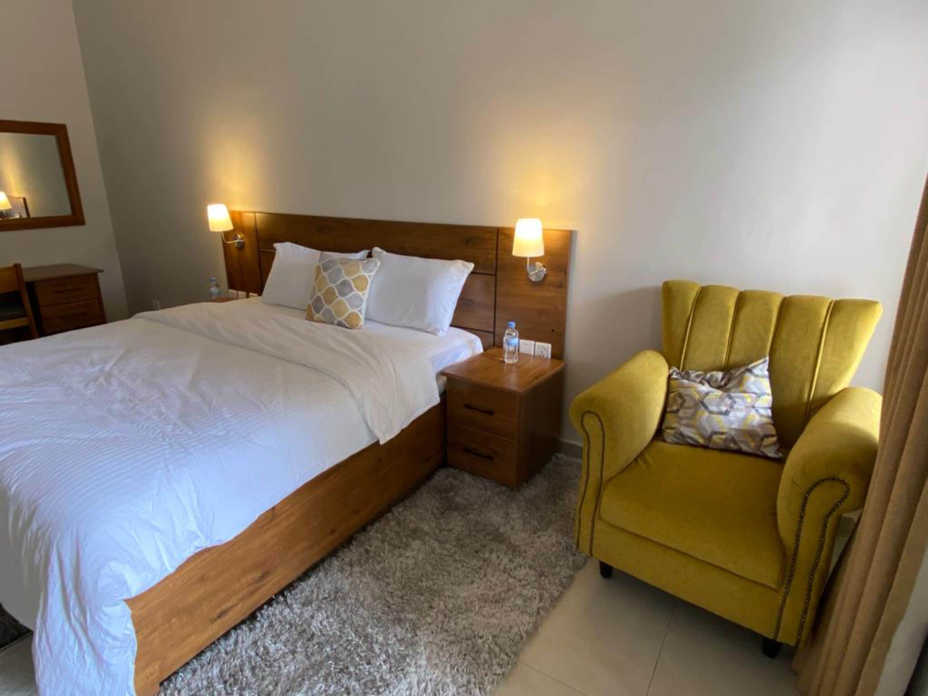 A hotel room with a large bed, a wooden headboard, two bedside tables with lamps, a yellow armchair, and a small table with a water bottle.