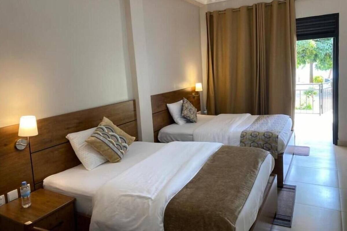 Une chambre d’hôtel avec deux lits, une table de chevet avec une lampe, une fenêtre avec des rideaux et une vue sur la verdure.