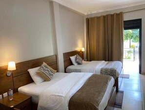 A hotel room with two beds, a nightstand with a lamp, a window with curtains, and a view of greenery outside.