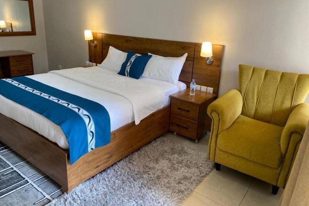 Un lit bien fait, recouvert d’une couvre-lit bleue et blanche, avec une tête de lit en bois, deux lampes de chevet, un fauteuil jaune et une petite table de chevet en bois sur laquelle se trouve une bouteille d’eau.