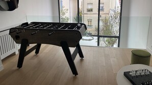 Ein Zimmer mit Kicker, Schachspiel und Blick auf einen Balkon mit Gebäuden und Bäumen.