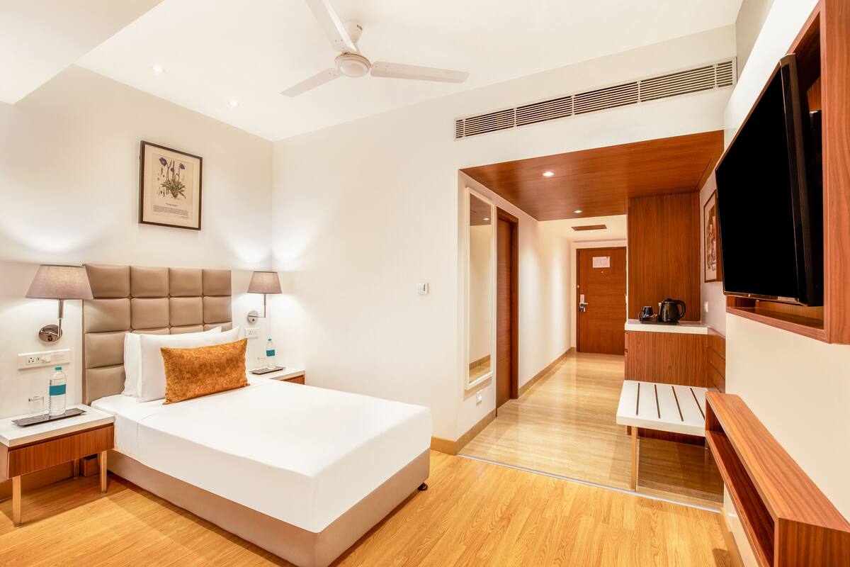 A modern hotel room with a large bed, bedside lamps, a ceiling fan, a TV, and a wooden bench.