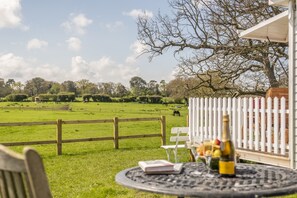 A table with a book and a bottle of champagne, a white fence, and a view of a green field with animals. - The Cricket Pavilion, Warham, Norfolk (Warham)