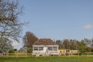 A small, single-story house with a white exterior and a brown roof, surrounded by a wooden fence and a grassy field. - The Cricket Pavilion, Warham, Norfolk (Warham)