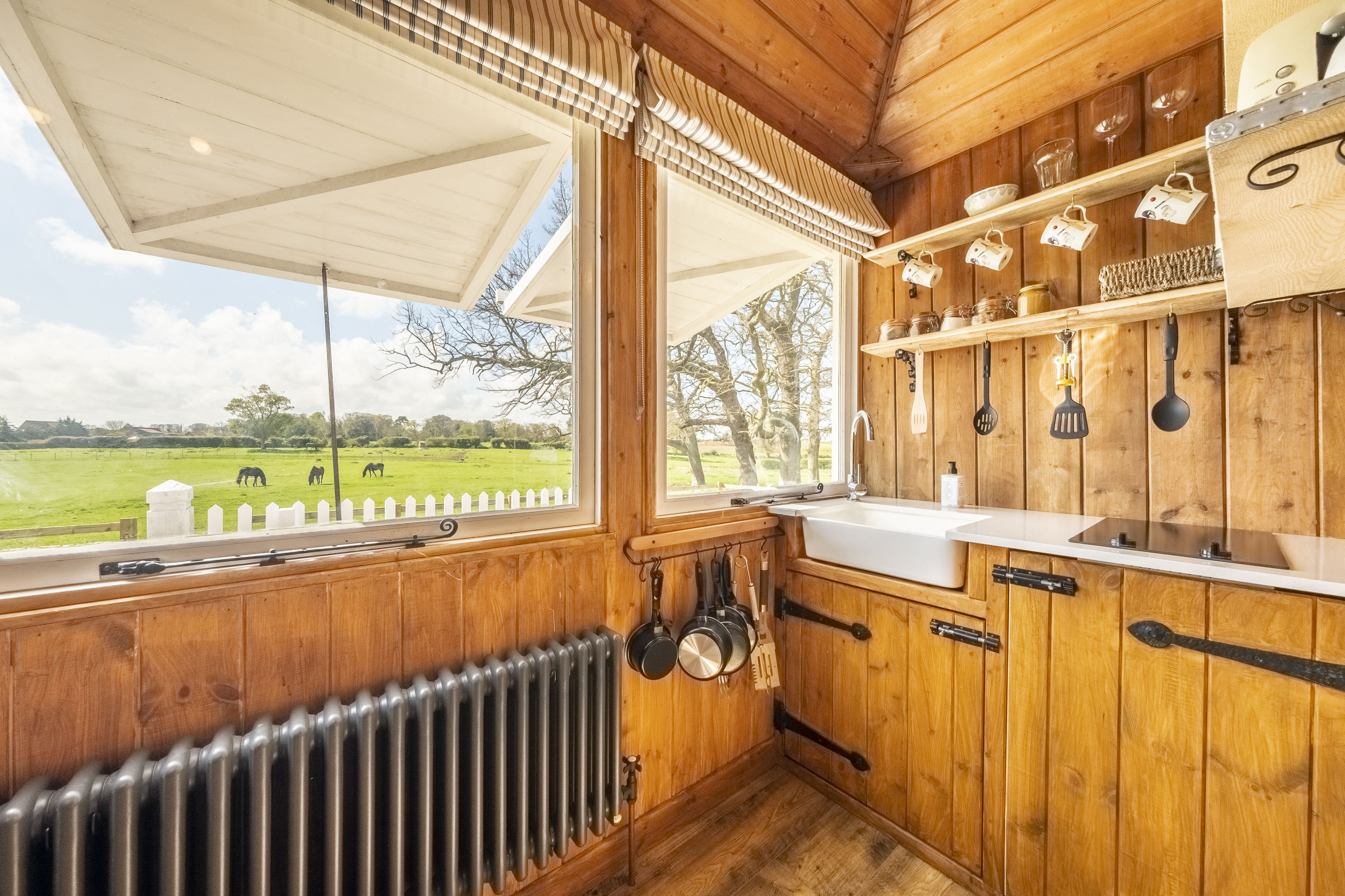A wooden interior with a large window, a kitchen area with utensils, and a view of a green field.