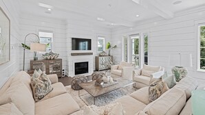 A spacious living room with a fireplace, beige sofas, and a large TV. - Royal Blue | Gated Neighborhood, Dedicated Beach Access, Golf Cart + Bikes (SANTA ROSA BEACH)