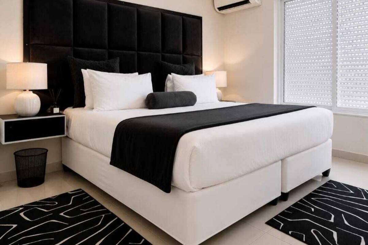 Una habitación de hotel moderna con una cama grande, dos mesitas de noche con lámparas, un cabecero negro y una ventana con persianas.