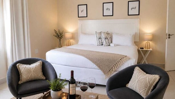 Une chambre à coucher avec un grand lit, deux fauteuils, une table de chevet avec une bouteille et des verres, et des œuvres d’art accrochées au mur.