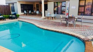 Une piscine dont le bord est revêtu de carrelage, entourée d’un espace de détente extérieur et d’un patio couvert.