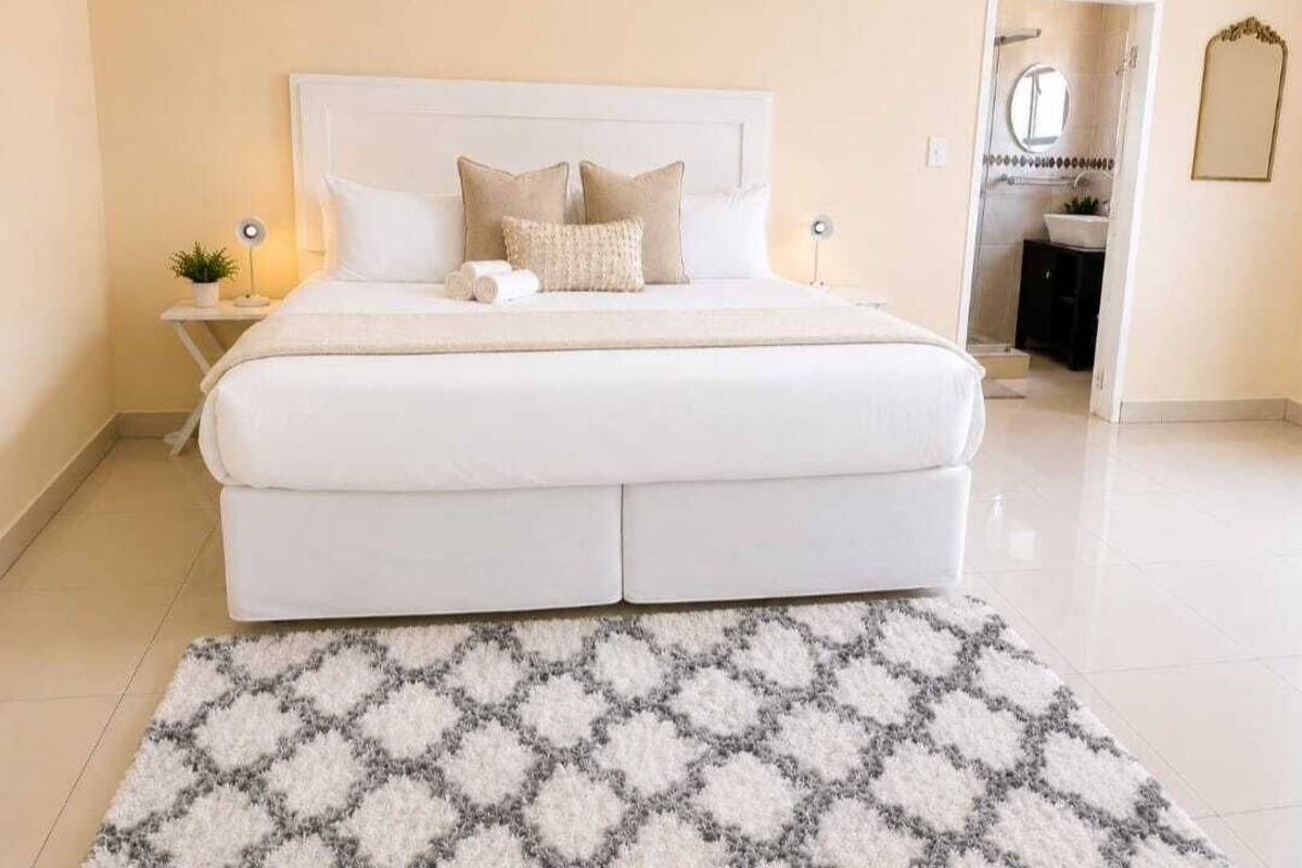 Una cama bien hecha con un cabecero blanco, una alfombra estampada y una mesita de noche con una lámpara.