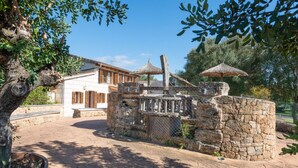 A stone gate with a wooden fence, a thatched roof structure, and a building with white walls and wooden shutters in the background. - Alcudia's Best: Private Pool, BBQ, Inviting Garden (Alcúdia)