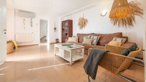 A spacious living area with a brown sofa, white coffee table, and wicker furniture. There is a hanging light fixture and a radiator on the wall. - Alcudia's Best: Private Pool, BBQ, Inviting Garden (Alcúdia)