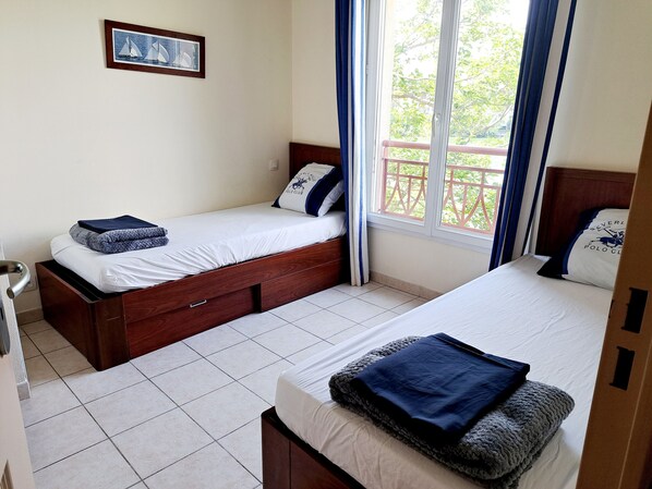 Een hotelkamer met twee eenpersoonsbedden, een houten hoofdbord, een raam met gordijnen, een lijst met een afbeelding aan de muur en een betegelde vloer.