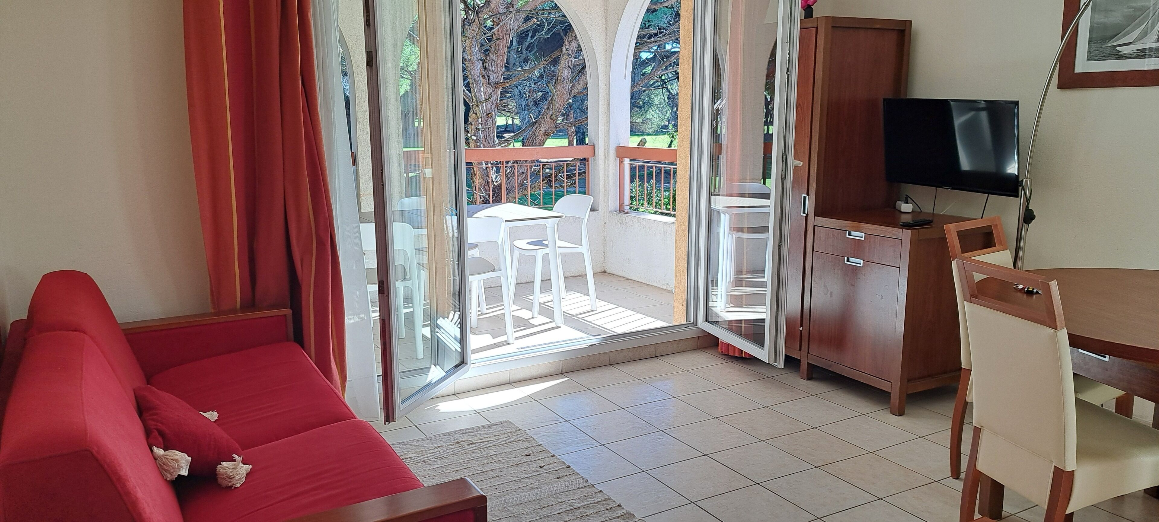 A room with a red sofa, a wooden desk, a television, and a sliding glass door leading to an outdoor area with a table and chairs.