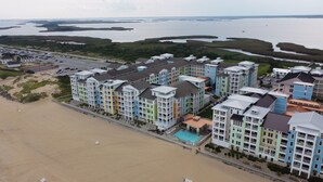 A coastal hotel with multiple colorful buildings, a beach, and a large body of water in the background. - Book Mermaid's Lookout 410B | Sandbridge Oceanfront Rental (Virginia Beach)