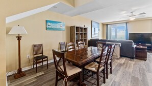 A dining area with a wooden table and chairs, a bookshelf, and a television in the living room. - Book Mermaid's Lookout 410B | Sandbridge Oceanfront Rental (Virginia Beach)