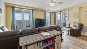 A spacious living room with a large sofa, a television, and a ceiling fan. - Book Mermaid's Lookout 410B | Sandbridge Oceanfront Rental (Virginia Beach)