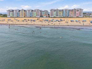 Et strandhotel med flere farverige bygninger, en sandstrand med parasoller og et roligt hav.