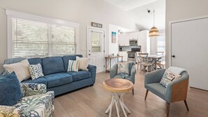 A living room with a blue sofa, a wicker armchair, and a wooden coffee table. There is a ceiling fan, a dining area with a table and chairs, and a kitchen in the background. - Heaven At The Sound: Fully Remodeled | Dogs OK (Kill Devil Hills)