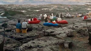 Une plage rocheuse où des personnes sont assises sur des tapis colorés, face à l’océan, au coucher du soleil.