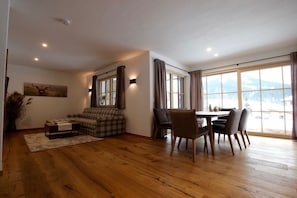 Un salon spacieux doté d’un plancher en bois, d’un canapé, d’une table à manger avec des chaises et de grandes fenêtres donnant sur l’extérieur.