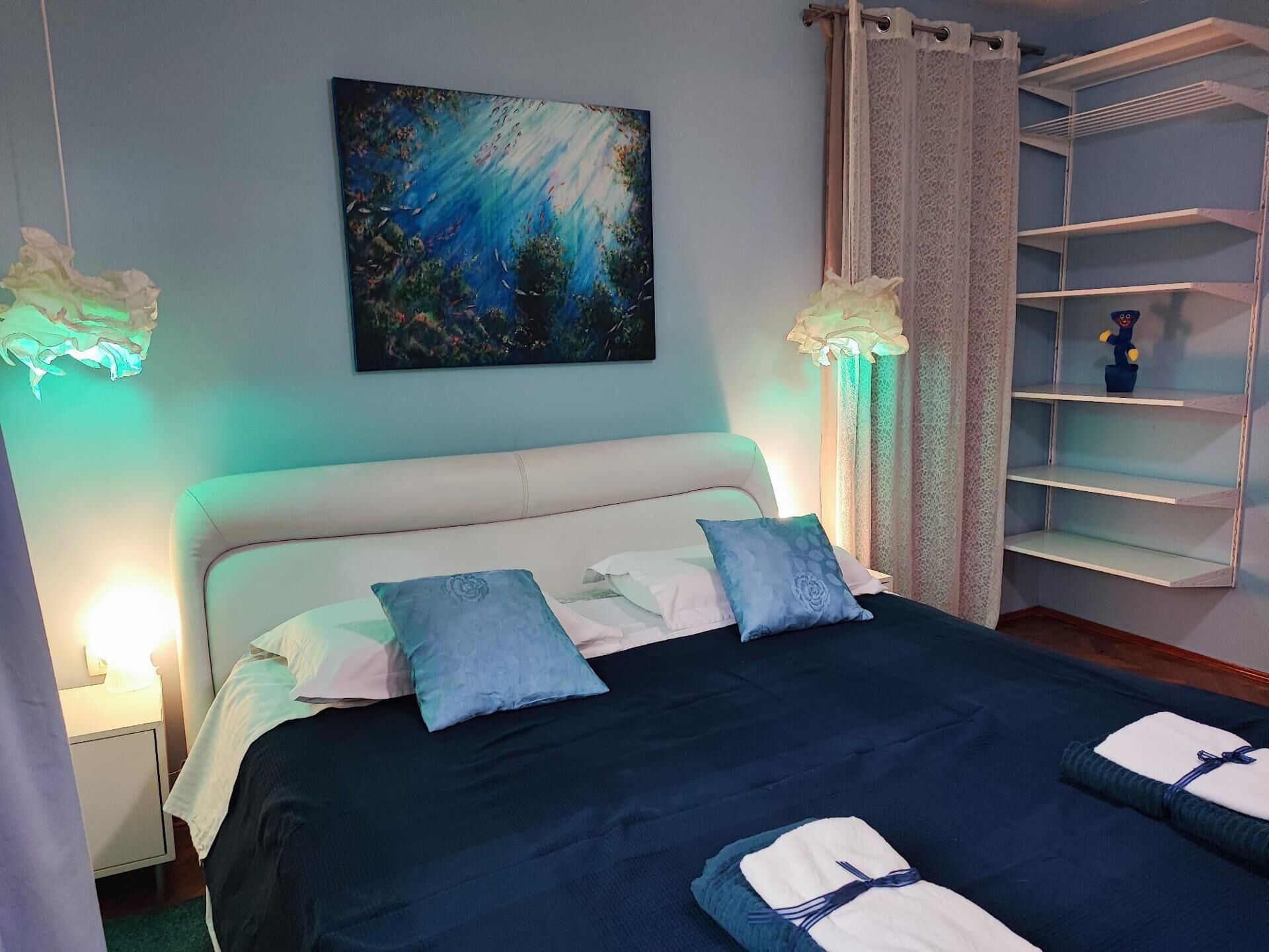 A bedroom with a bed, pillows, a painting, a nightstand, a lamp, and a shelf.