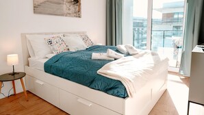 Une chambre à coucher avec un lit, une table de chevet, une lampe, un tableau et une vue sur un immeuble par la fenêtre.