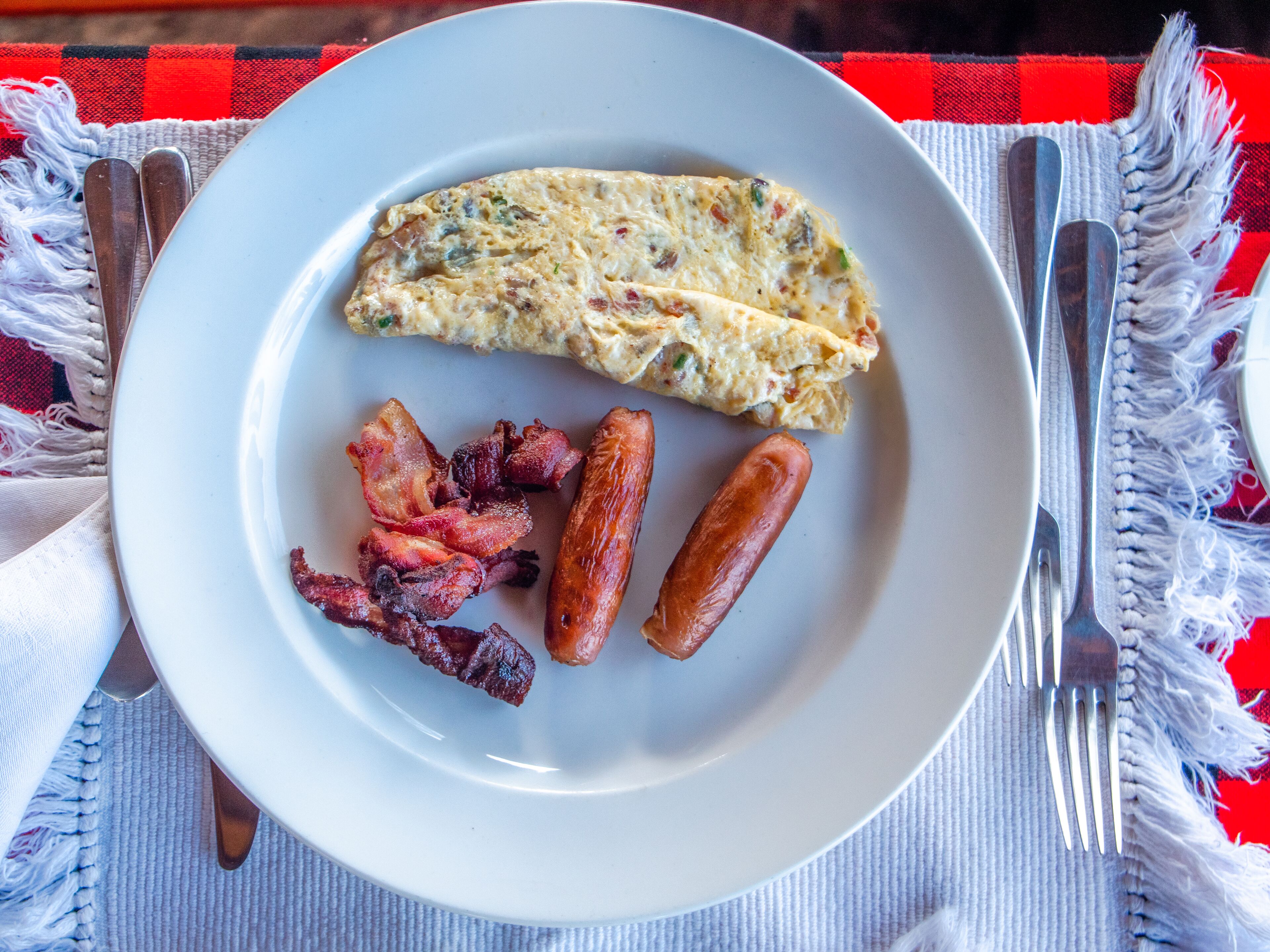 A plate with an omelette, bacon, and sausages on a tablecloth.
