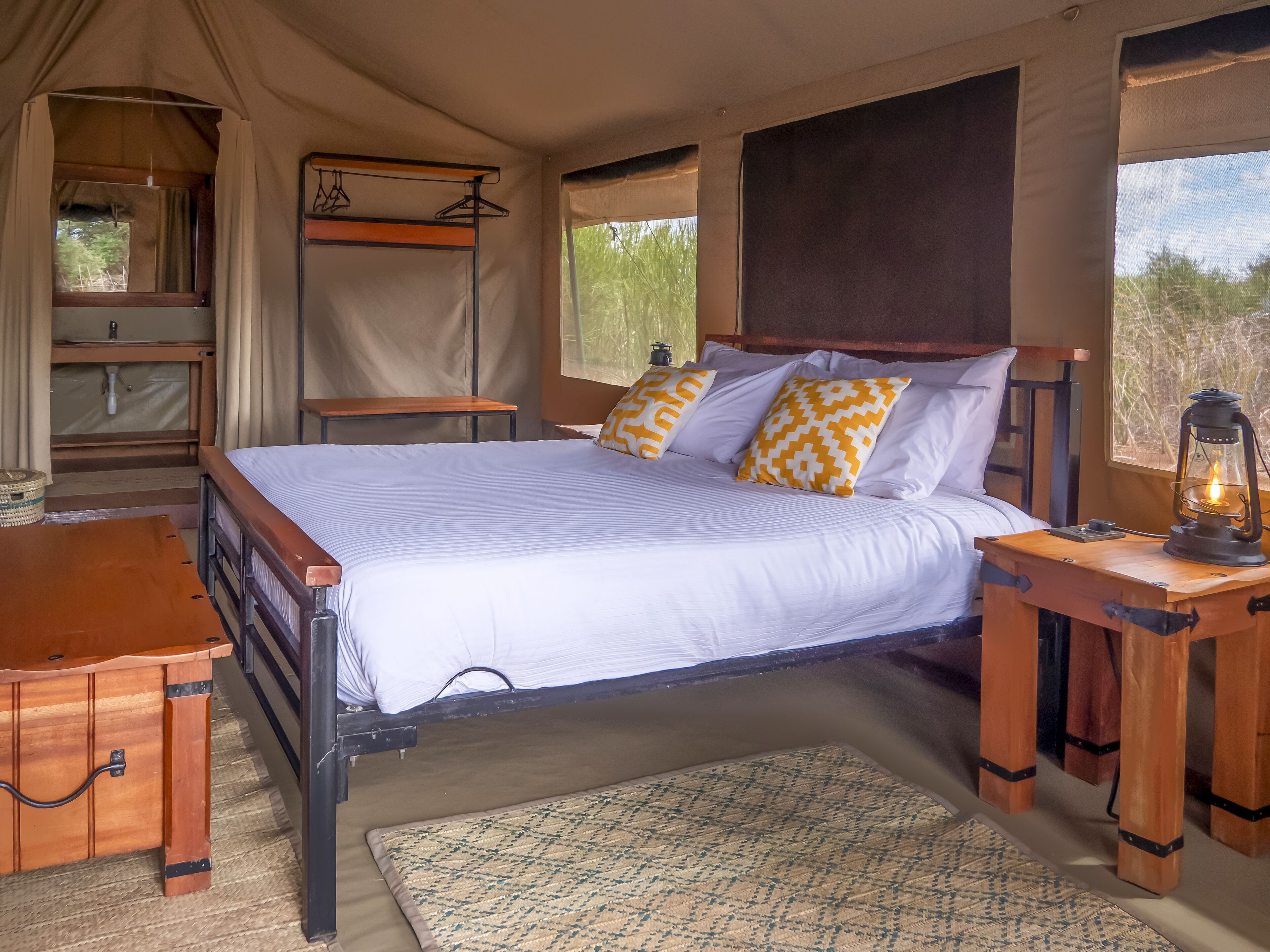 A bed with white bedding and patterned pillows, a wooden bedside table with a lantern, a small wooden desk, a hanging rack with clothes, and a window with a view of trees.
