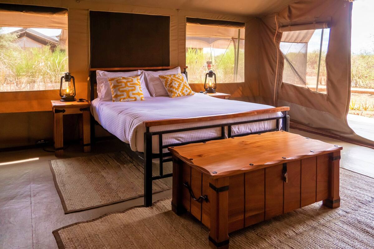 A tented room with a bed, two bedside tables, a wooden bench, and a view of the outdoors.
