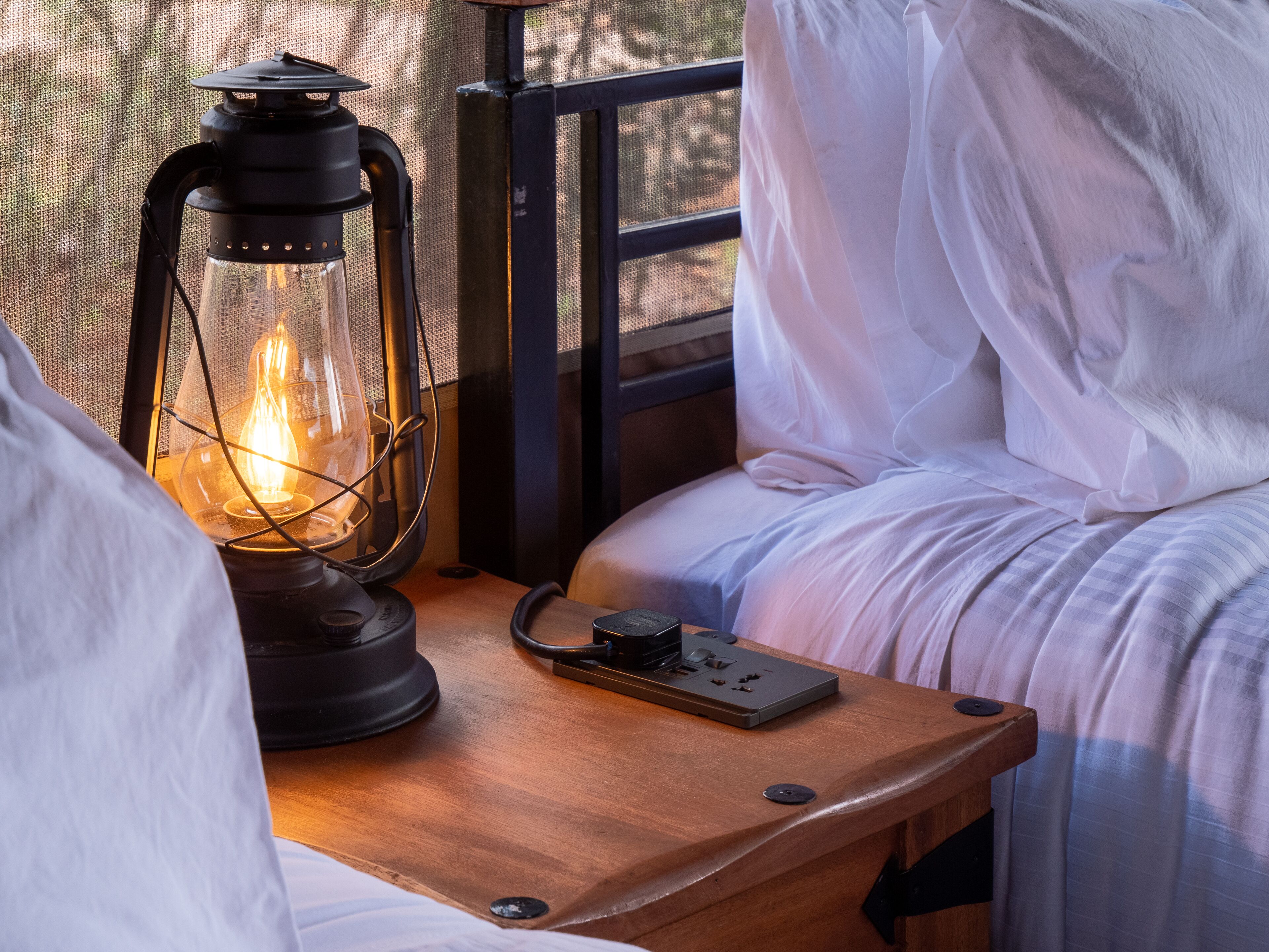 A bed with white bedding, a wooden nightstand, a lantern, and a power outlet.