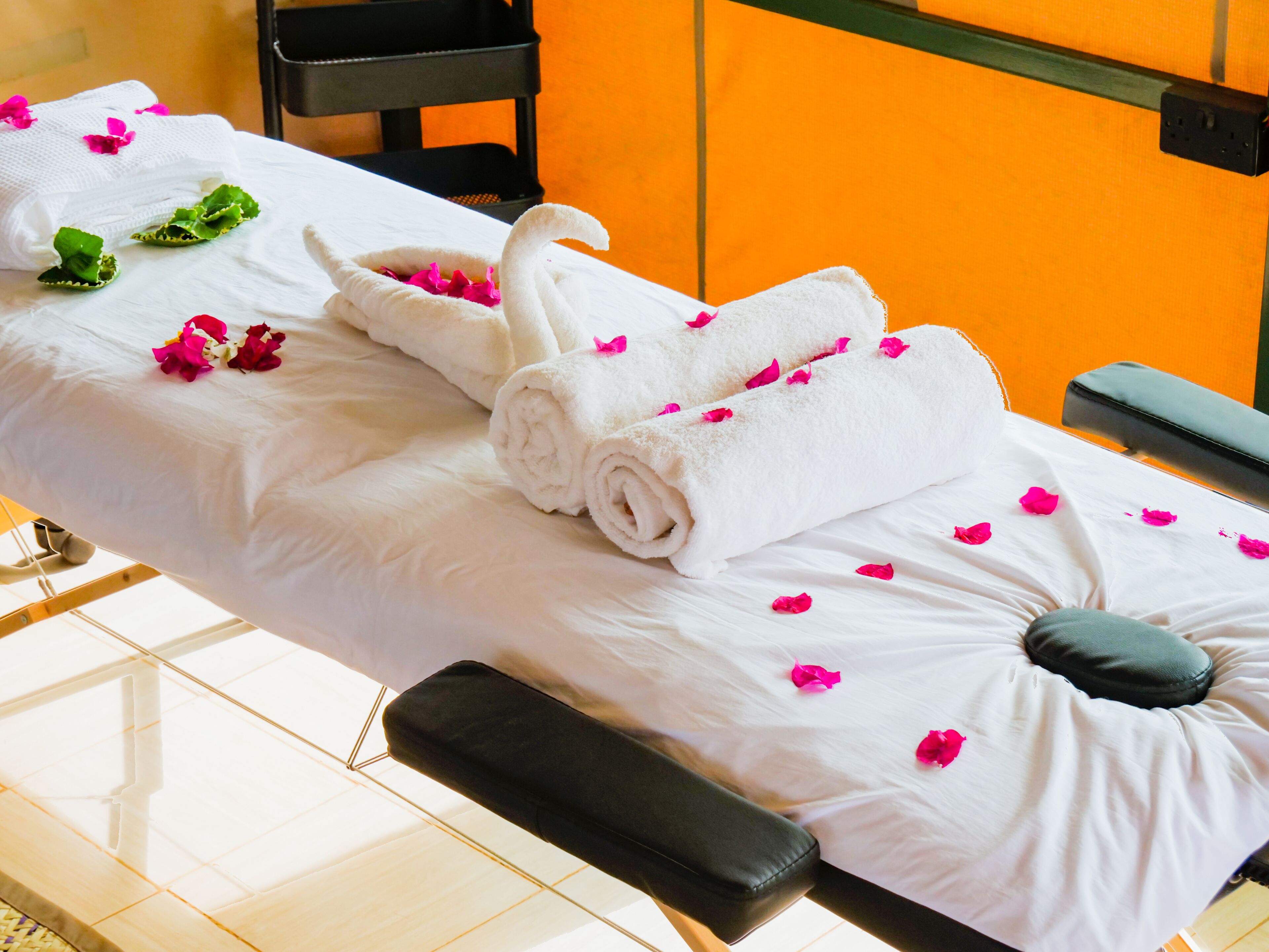 A massage table with rolled towels and pink flower petals, a black massage stool, and a black massage bench.