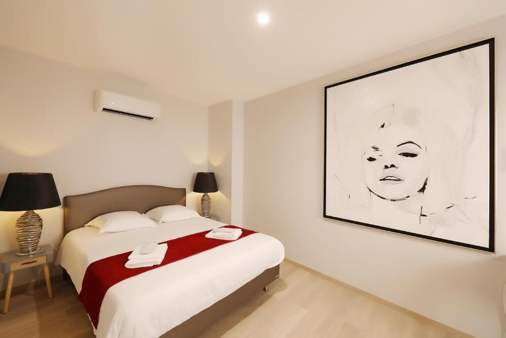 A hotel room with a bed, two bedside lamps, an air conditioner, and a large abstract painting on the wall.