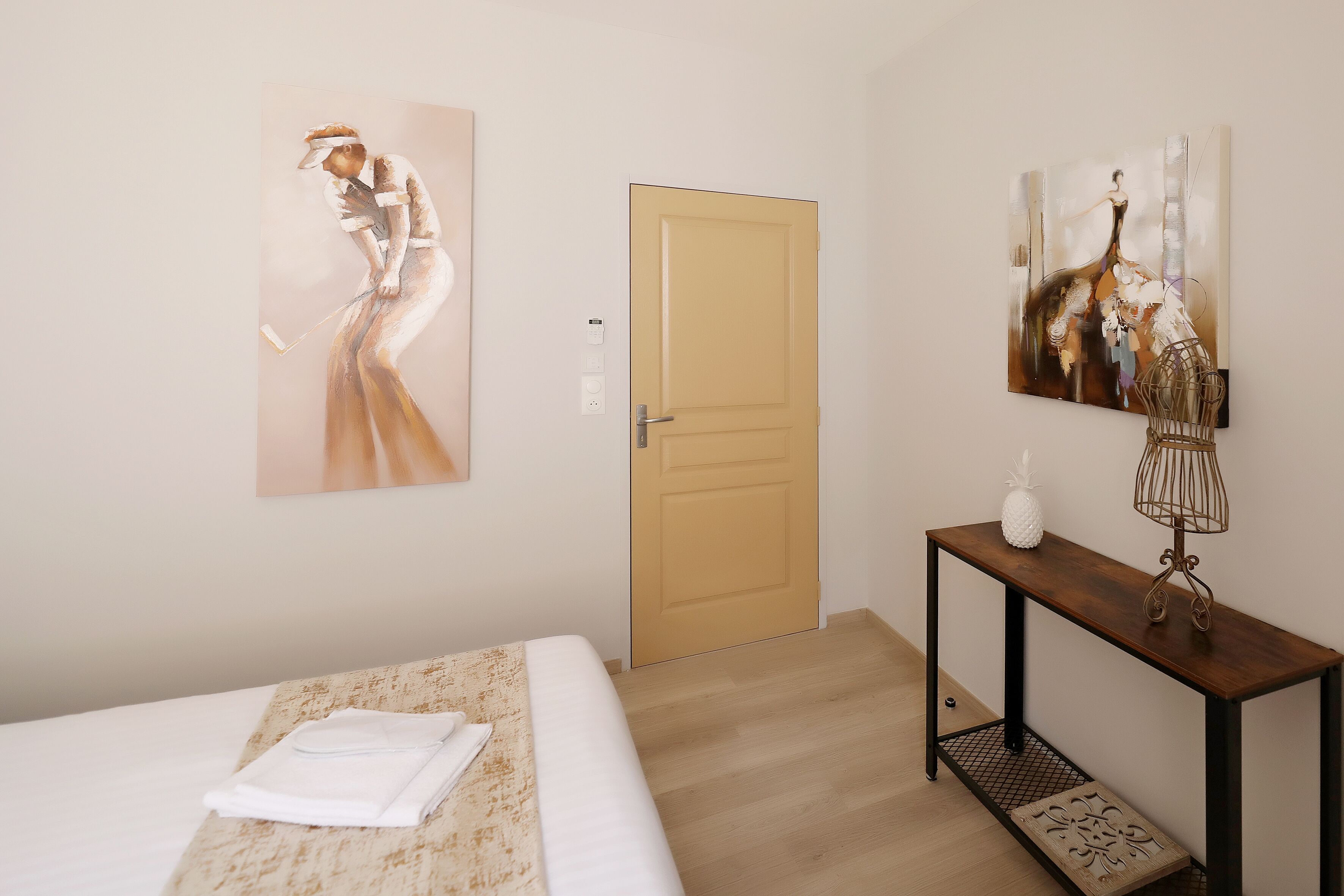 Une chambre d’hôtel avec un lit, une console en bois sur laquelle se trouve une lampe, et deux tableaux accrochés au mur.