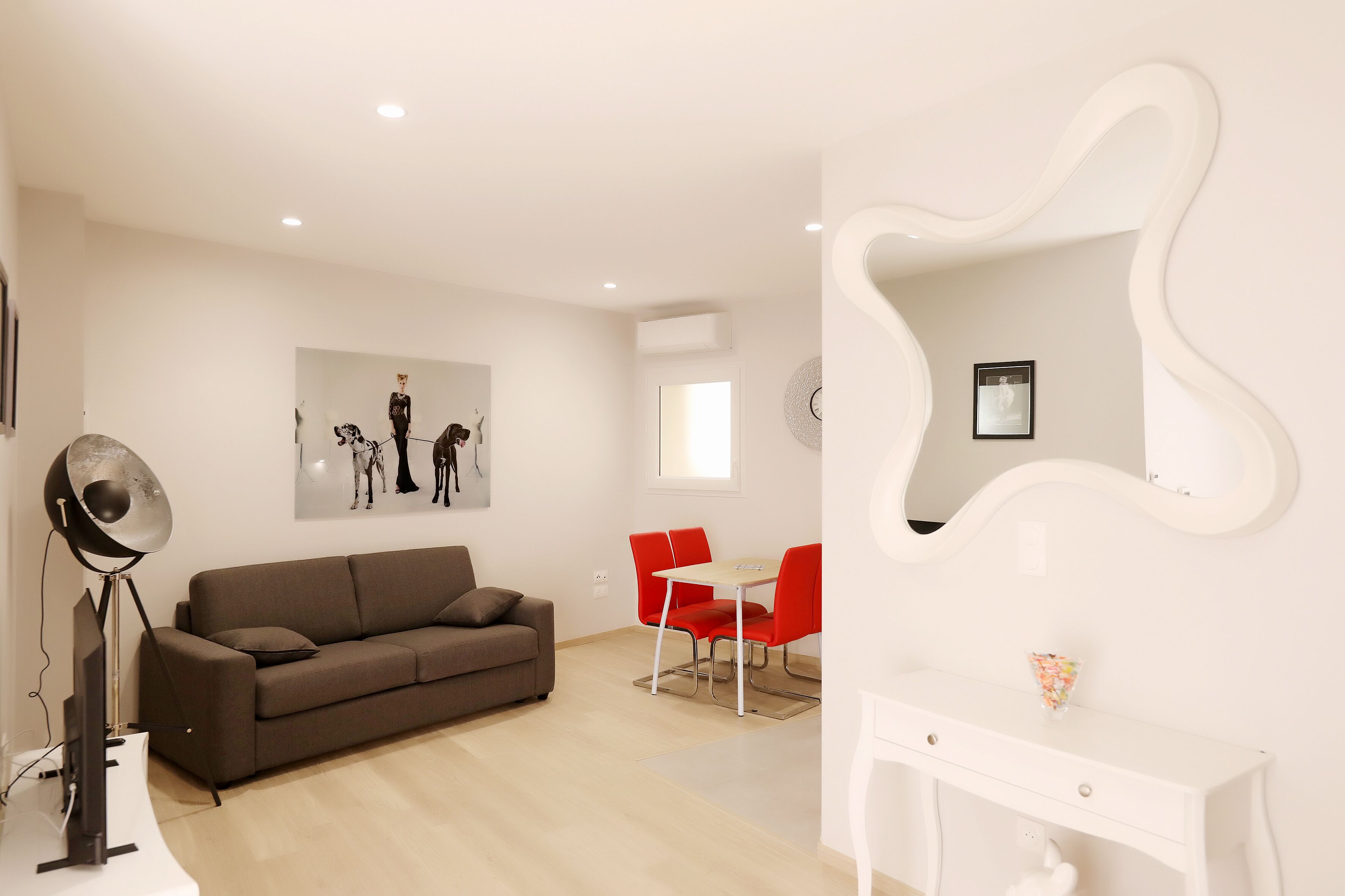 A modern living room with a sofa, a dining table with red chairs, a white console table, and a large mirror.