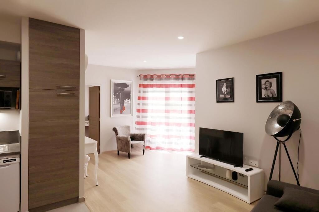 Una sala de estar moderna con un televisor de pantalla plana, un mueble blanco para entretenimiento y una lámpara de pie. Hay una zona de comedor con una mesa y sillas blancas, y una cocina con horno y microondas integrados.
