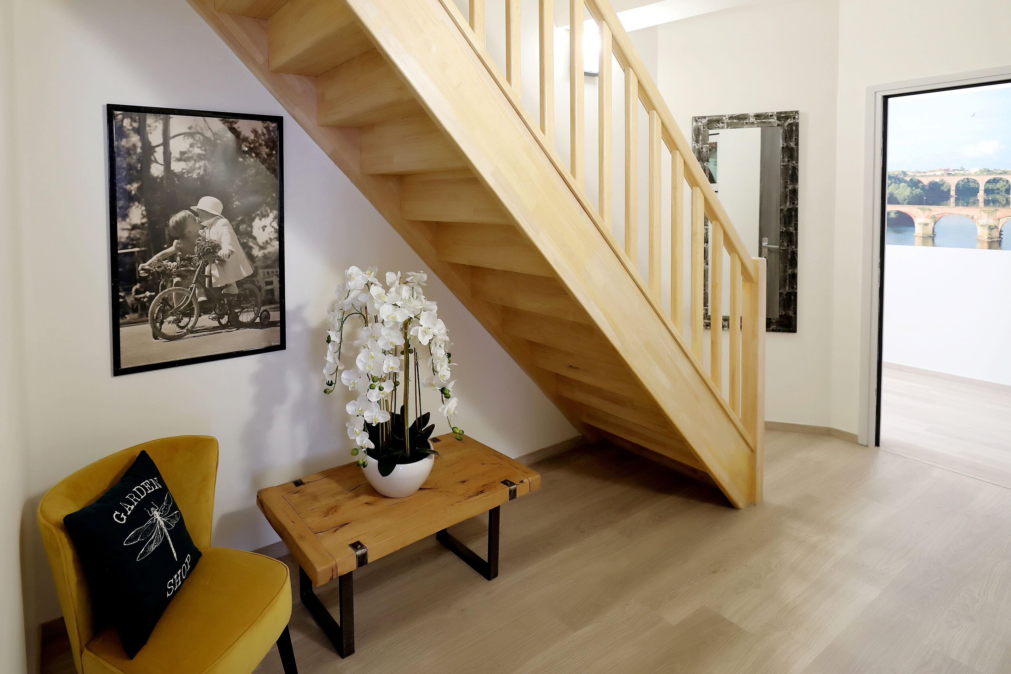 Un escalier avec des rampes en bois, une photographie en noir et blanc encadrée accrochée au mur, une chaise jaune avec un coussin noir et une petite table sur laquelle est posé un vase blanc rempli de fleurs.
