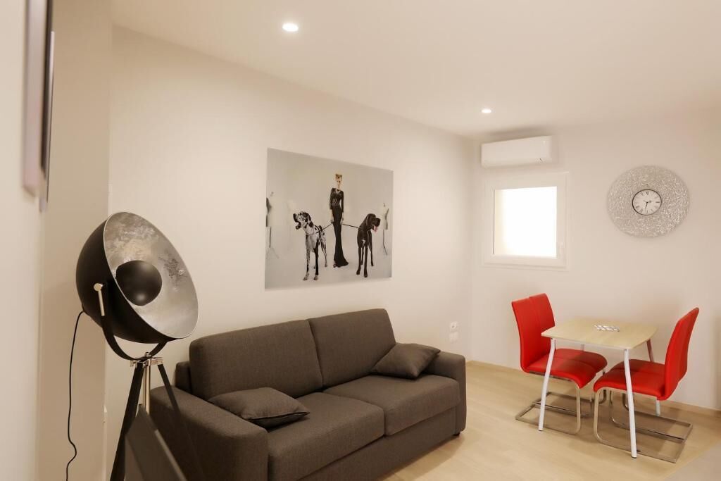 A living room with a grey sofa, a red dining table with two chairs, and a wall-mounted artwork.