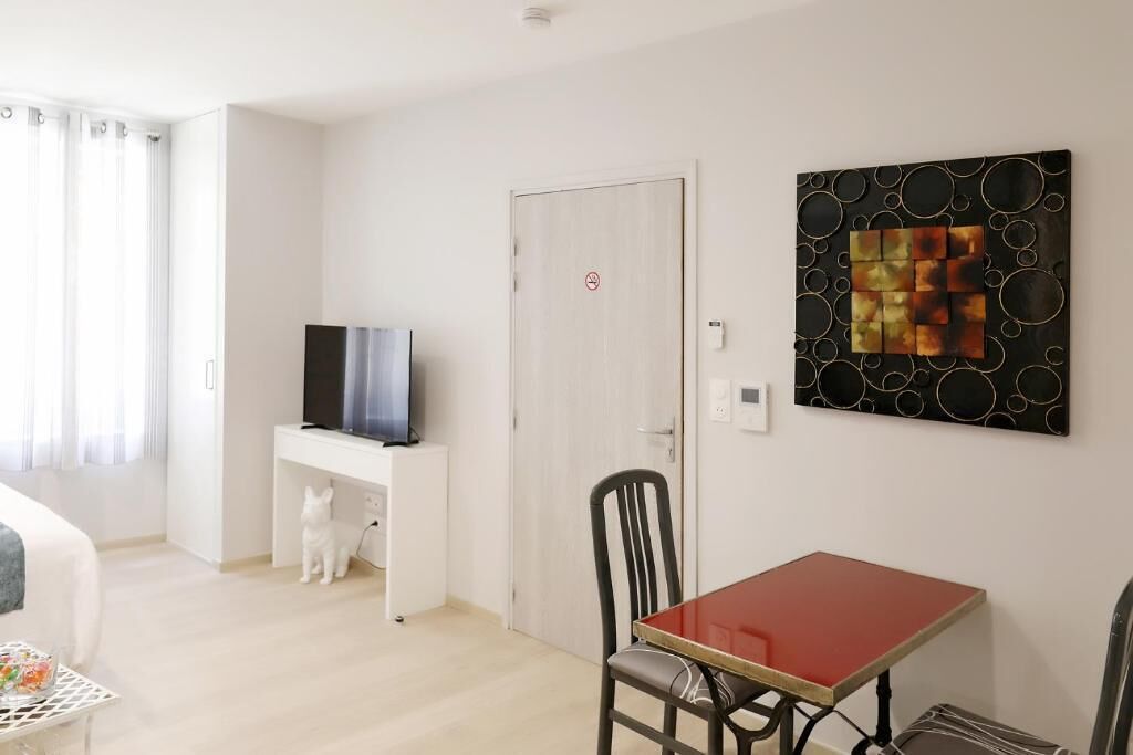 Een kamer met een bed, een bureau met een televisie, een eettafel met stoelen en een muur met een geometrisch kunstwerk.