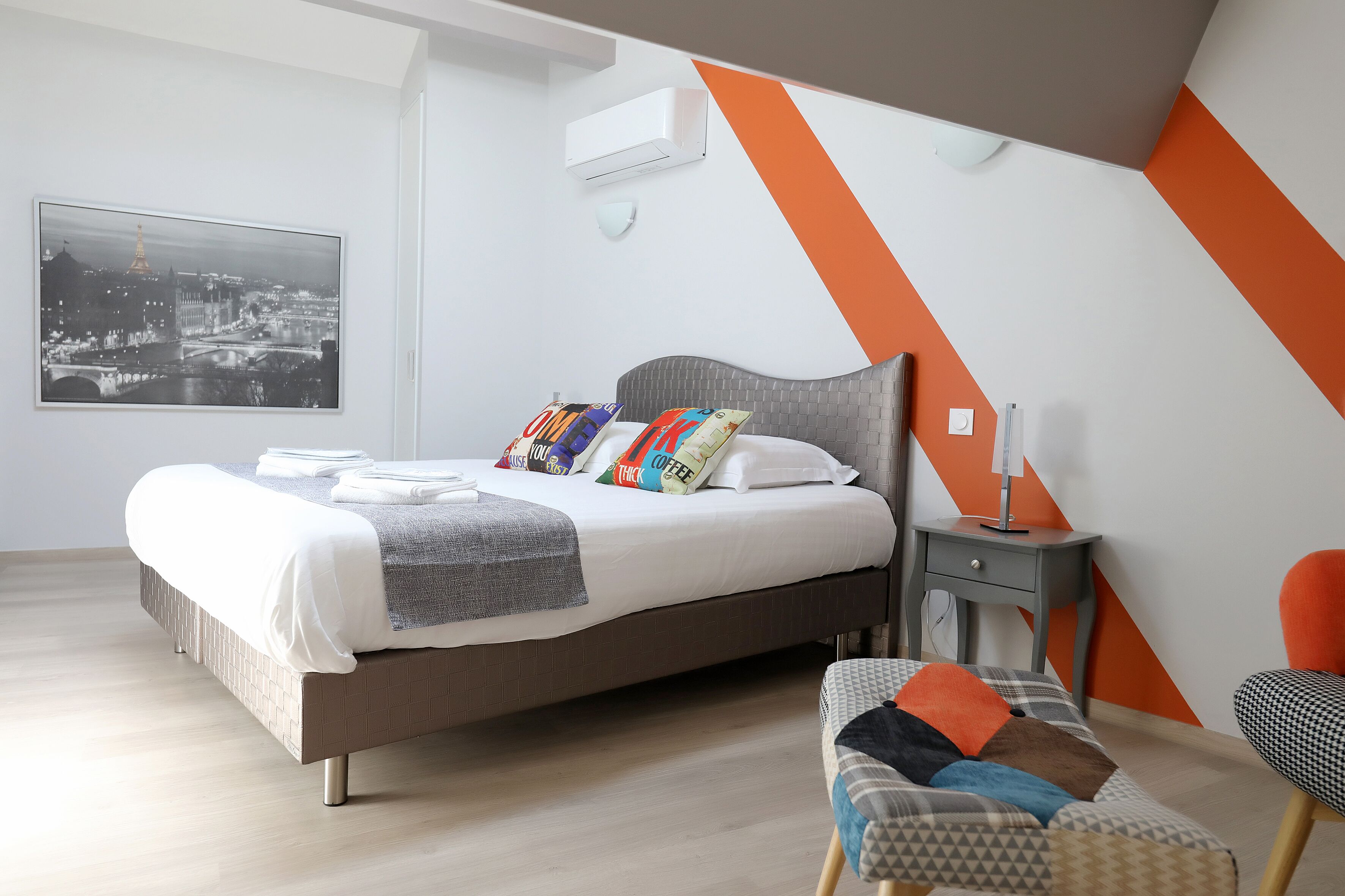 Um quarto moderno com uma cama grande, vista para a cidade, uma parede com detalhes em cinza e laranja e uma pequena mesa de cabeceira.