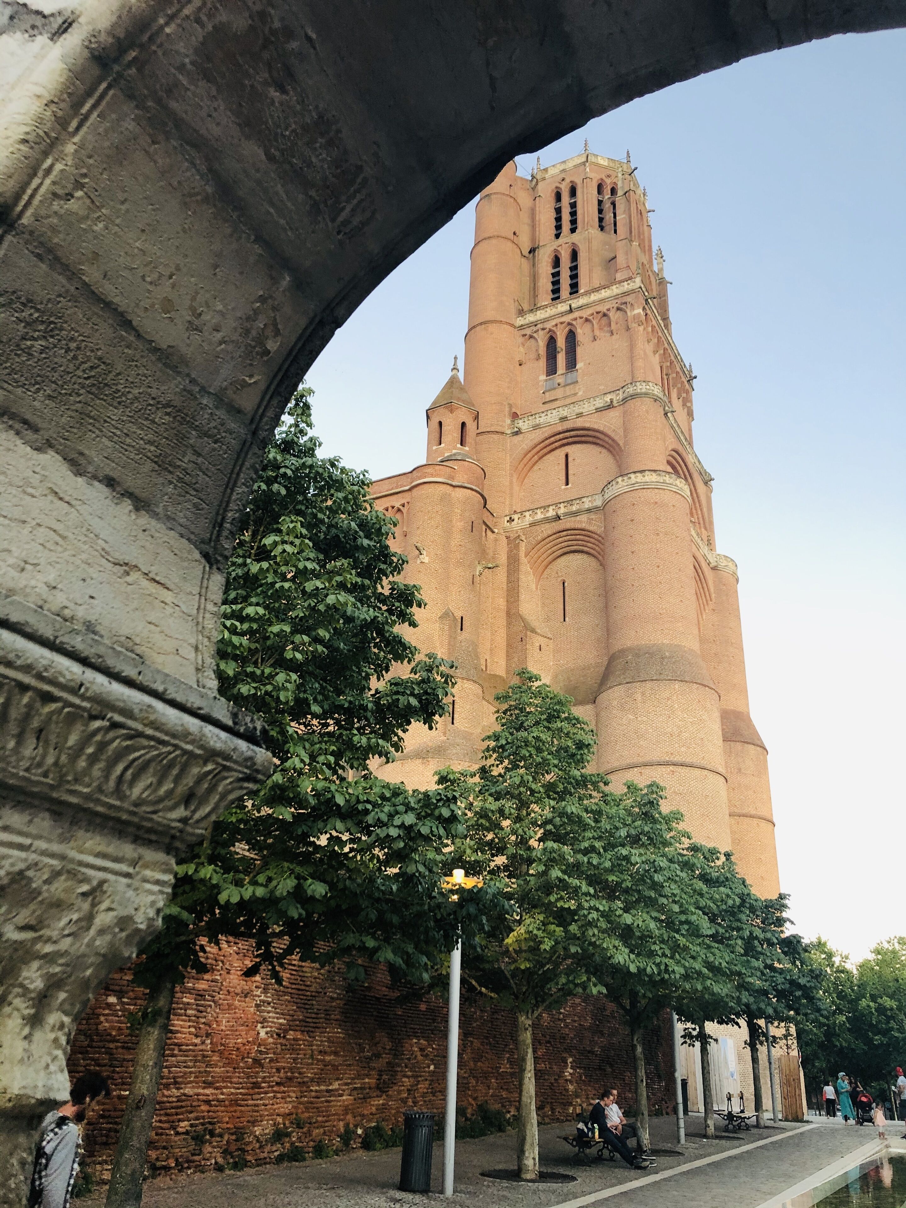 Un edificio histórico con una torre destacada, un arco de piedra y un paseo adoquinado con árboles y bancos.