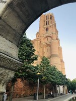 Un edificio histórico con una torre destacada, un arco de piedra y un paseo adoquinado con árboles y bancos.