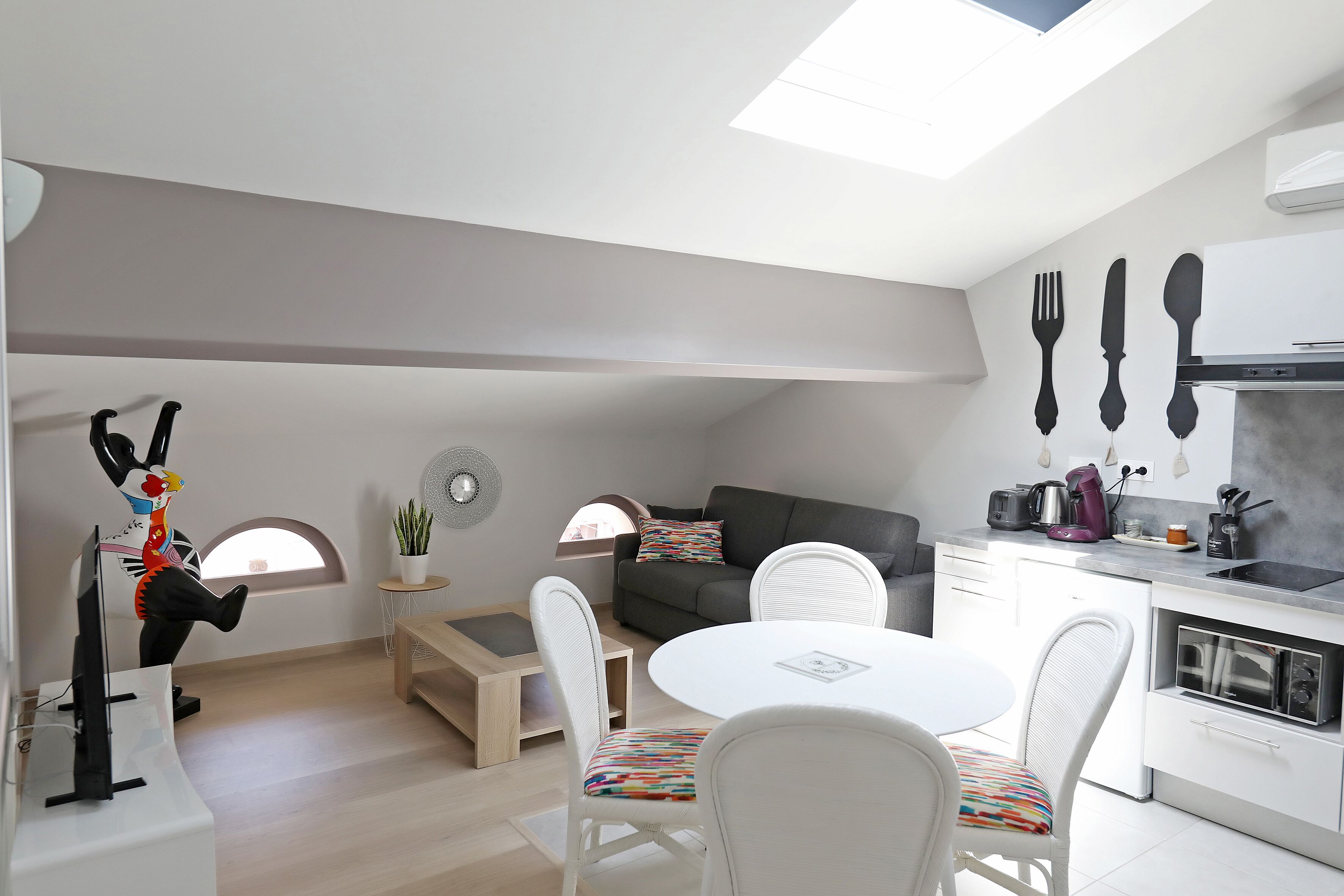 Eine moderne Küche mit weißen Schränken, ein Essbereich mit rundem Tisch und Stühlen, ein Sofa und ein kleiner Beistelltisch mit einer Pflanze.