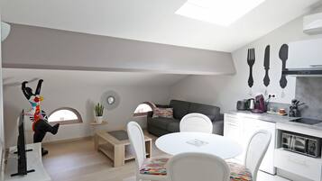Eine moderne Küche mit weißen Schränken, ein Essbereich mit rundem Tisch und Stühlen, ein Sofa und ein kleiner Beistelltisch mit einer Pflanze.