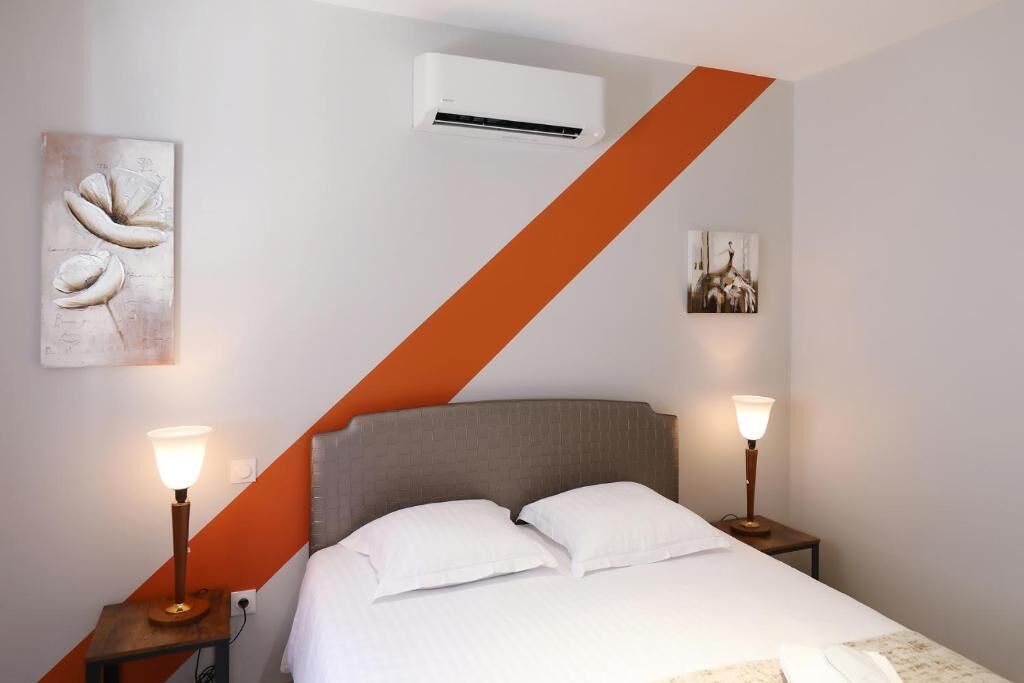 A hotel room with a bed, two bedside tables with lamps, an air conditioning unit, and artwork on the walls.