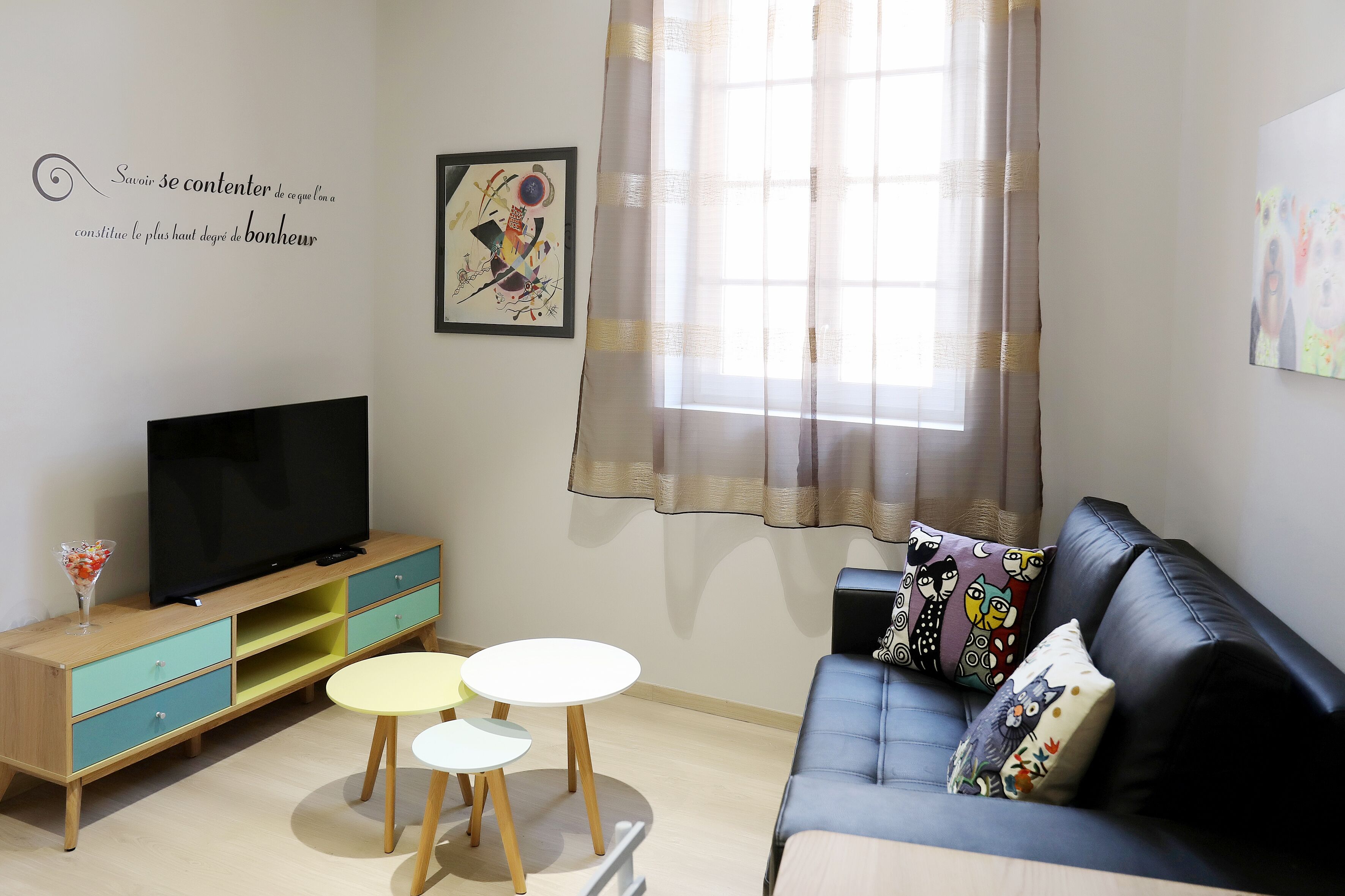 A living room with a TV on a wooden cabinet, a black sofa with patterned cushions, and a small white table.